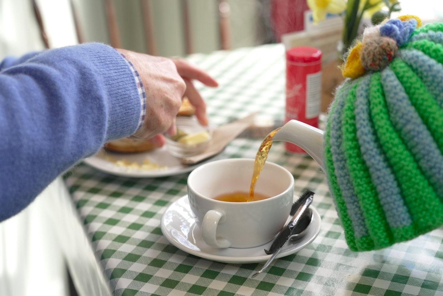 Tea is poured from a knitted teapot into a cup.
